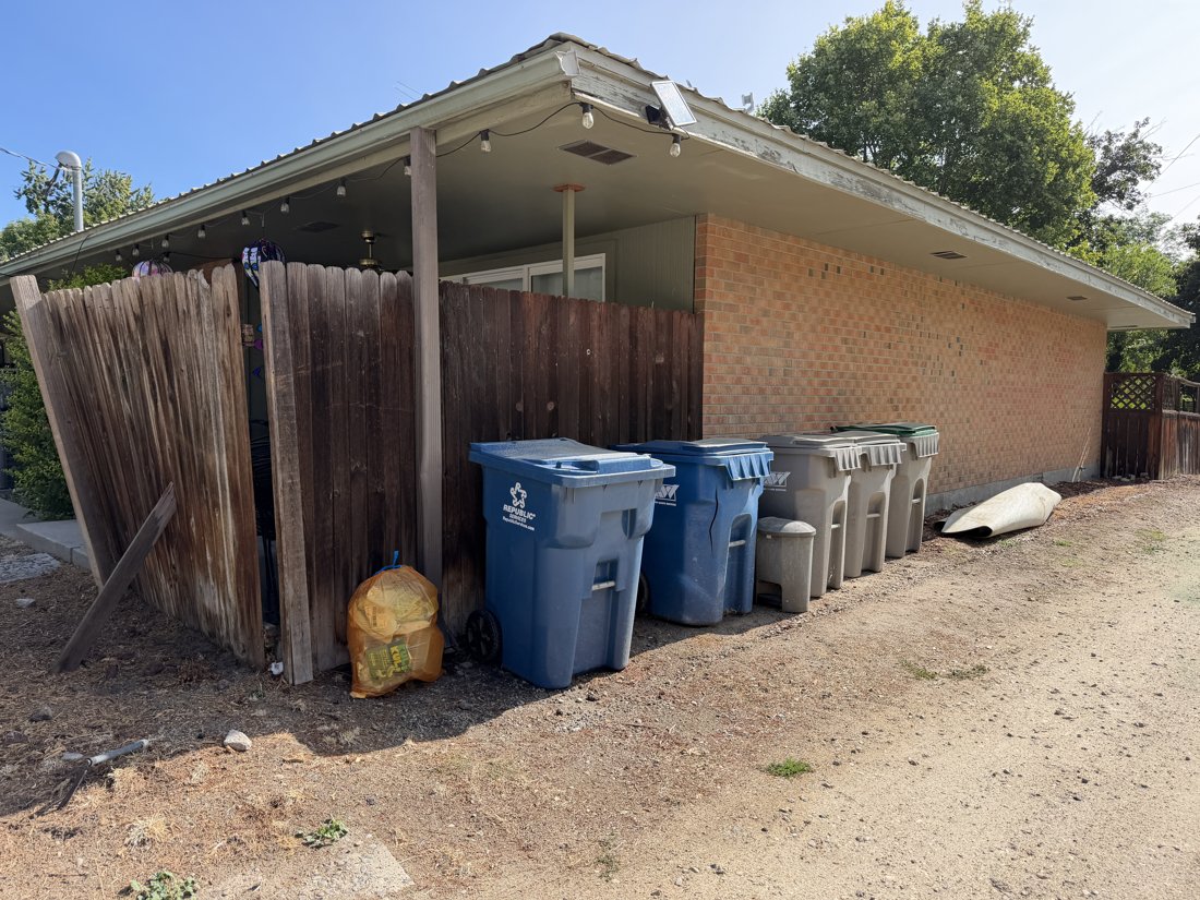 West side, viewed from the east. Alley service elevation with brick wall, deep eaves, fence, and refuse bins.