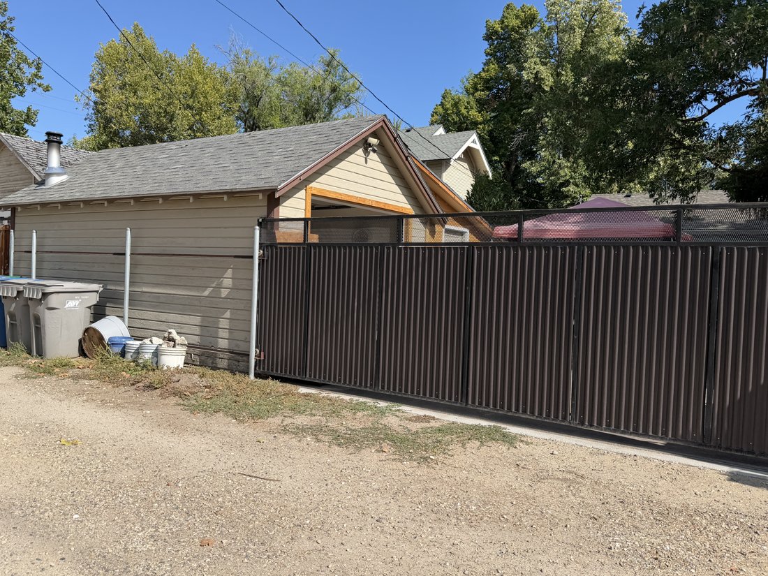 South side, viewed from the north. Rear outbuilding with a gabled asphalt-shingle roof, lap siding, exposed rafter ends, metal flue, and tall gate.
