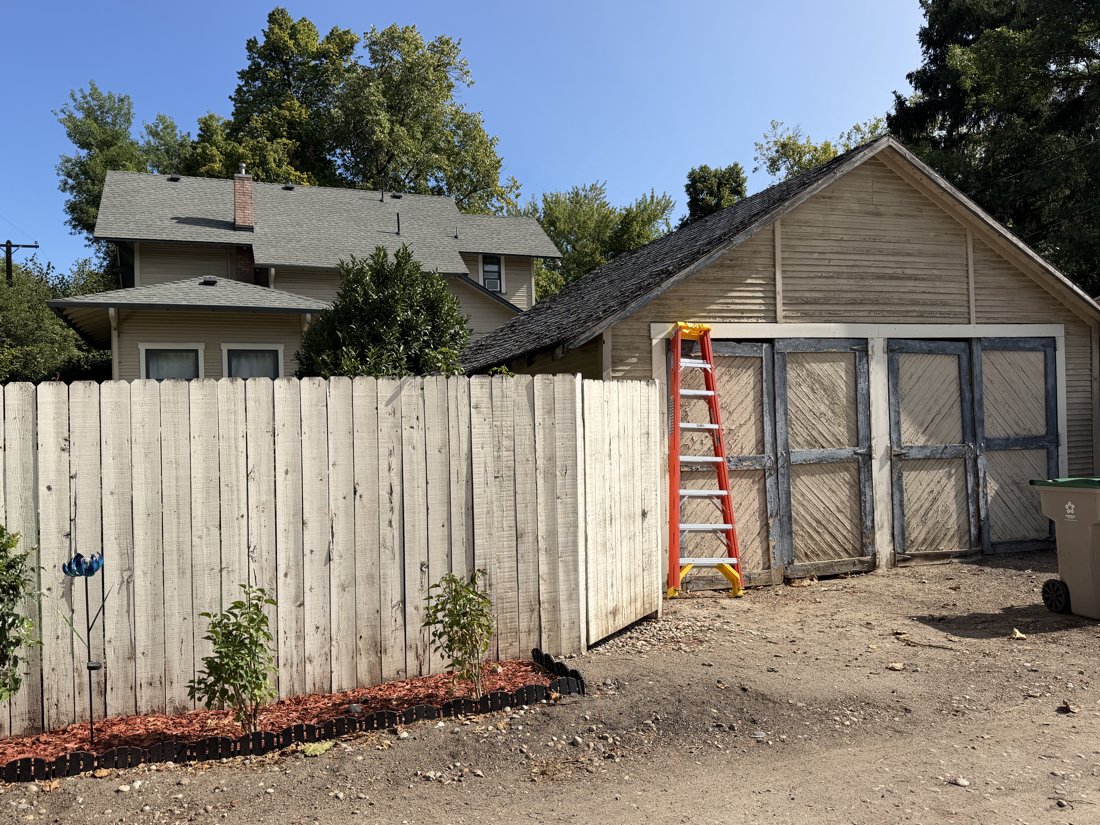 South and west sides, viewed from the northeast. Garage and rear of the house together; a low rear wing, rectangular windows, and brick chimney are visible beyond the fence.